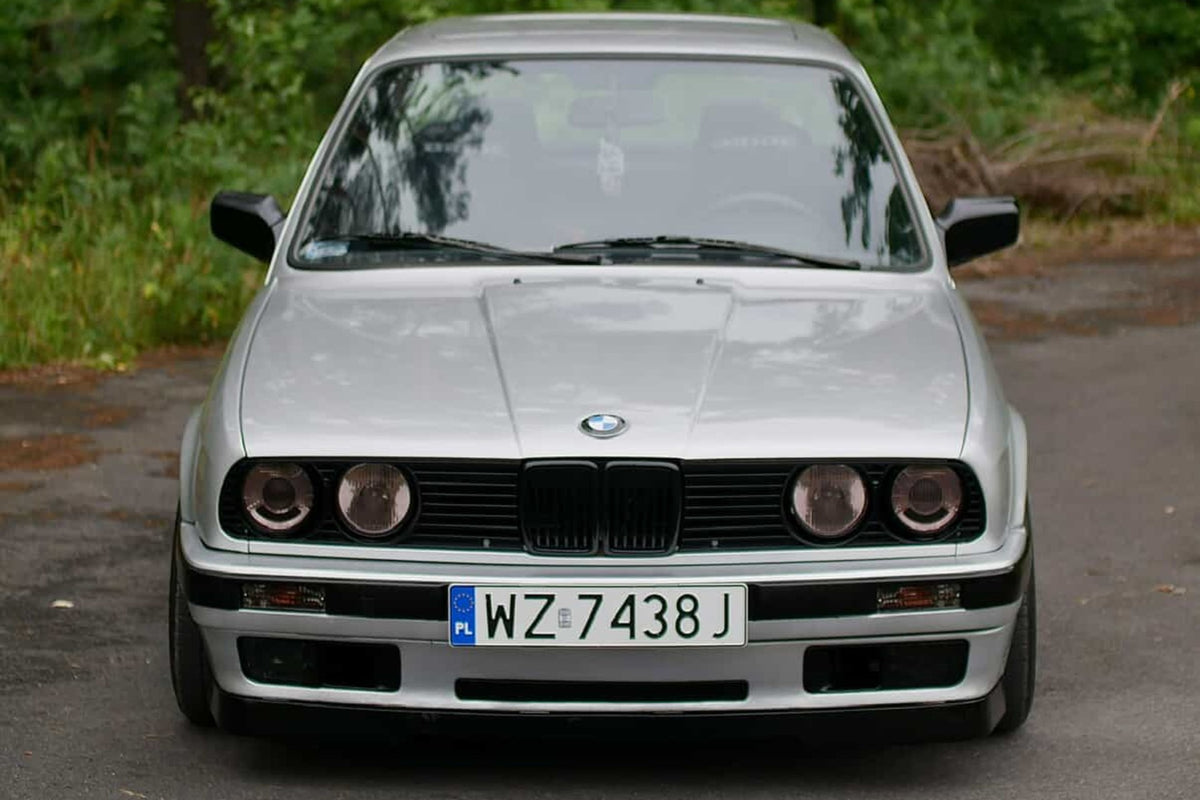 Szare bmw E30 od przodu z bezramkowymi mocowaniami tablic rejestracyjnych na tle lasu